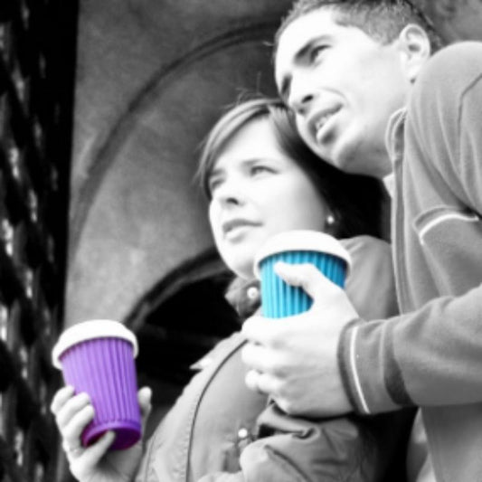 Two friends chat while holding Onya reusable coffee cups. One cup is purple with a white lid, the other cup is aqua blue with a white lid. Available at Green Distributors.