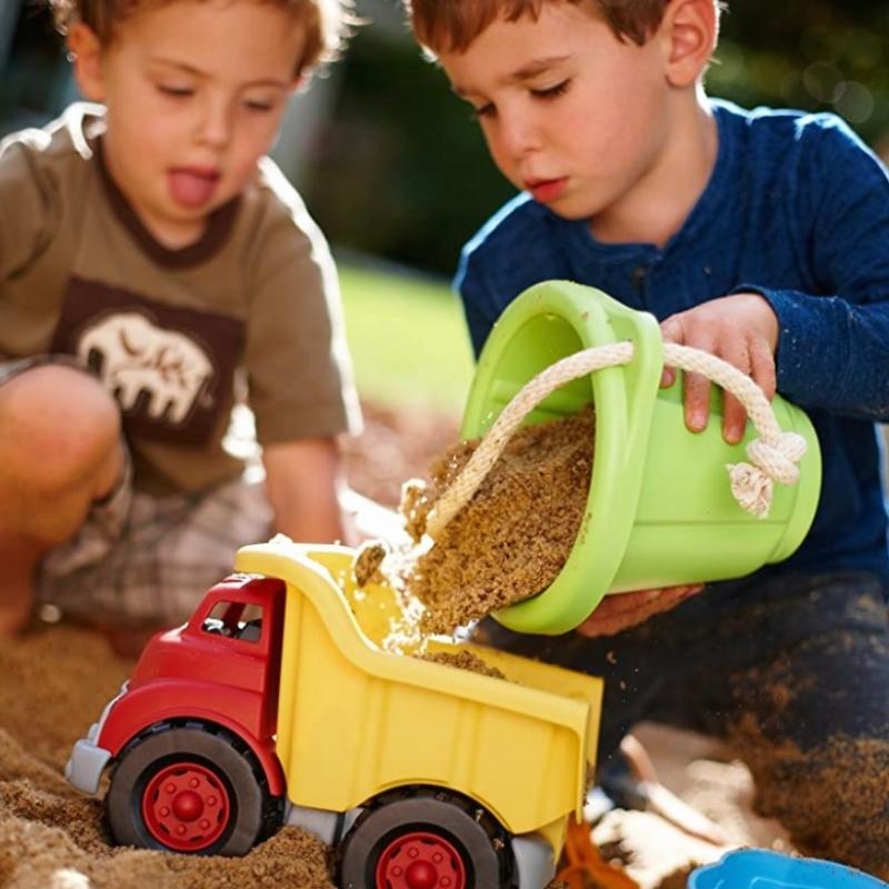 Children playing with Green Toys red dump truck in a sandbox. The kids are filling the back of the truck with sand from Green Toys Sand Play Bucket with a rope handle. Available at Green Distributors.