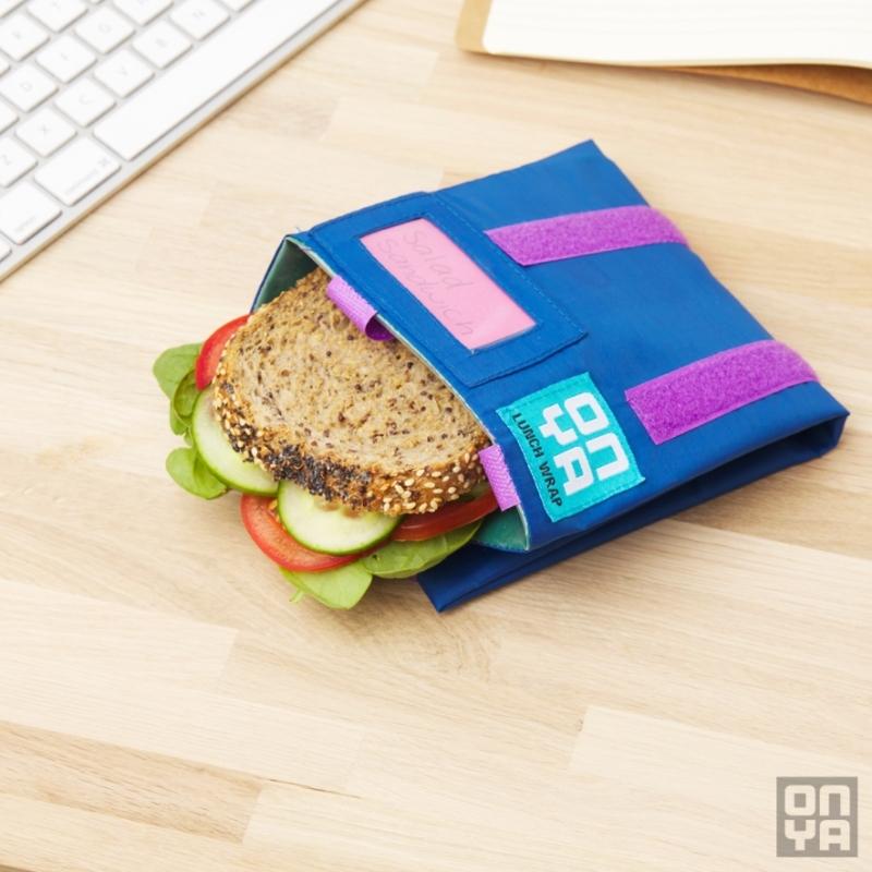 Lunch at a desk. A sandwich peaks out of an Indigo colored reusable lunch wrap at a desk with a keyboard. Available at Green Distributors.