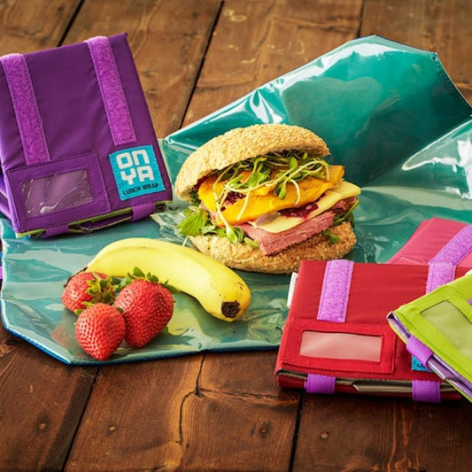 A sandwich, a banana, and some strawberries sit on an indigo colored adjustable size reusable lunch wrap by Onya. Pink, Purple, Red, and Apple colored wraps are set off to the side. Available at Green Distributors.