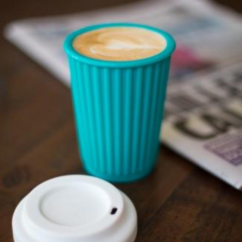 12 oz Onya reusable aqua colored coffee cup with artfully frothed java next to a white silicone lid and a newspaper. Available at Green Distributors.