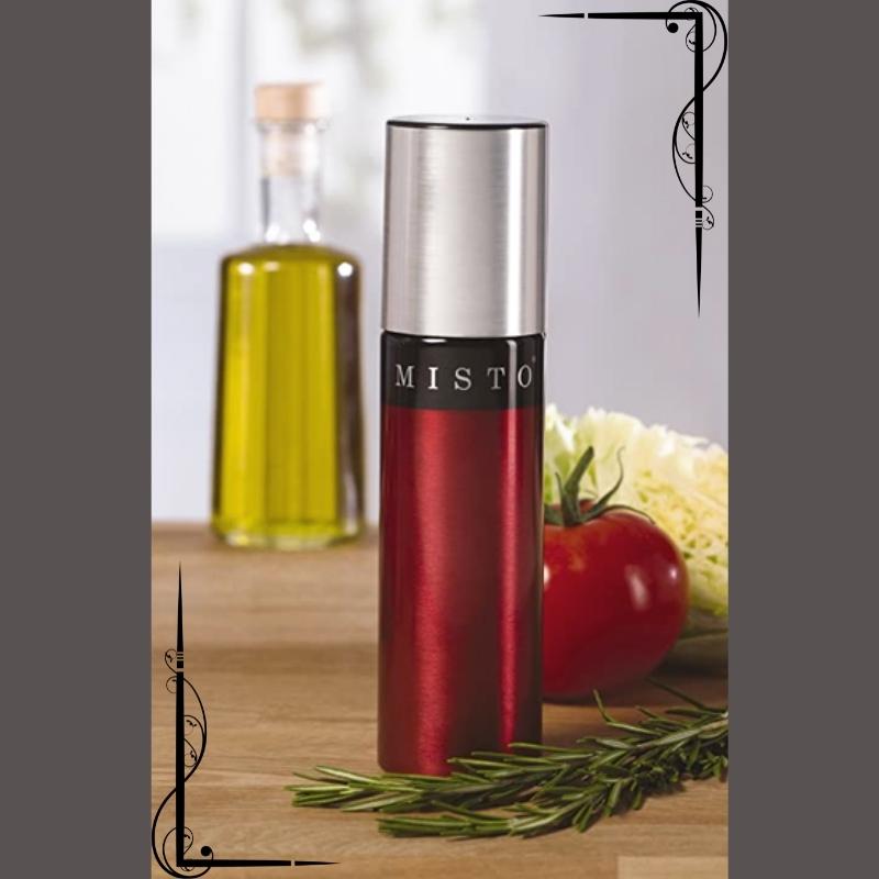 Tomato red Misto gourmet oil spraying bottle sits on a kitchen counter with a sprig of herbs, a tomato, and an oil bottle. Available at Green Distributors.